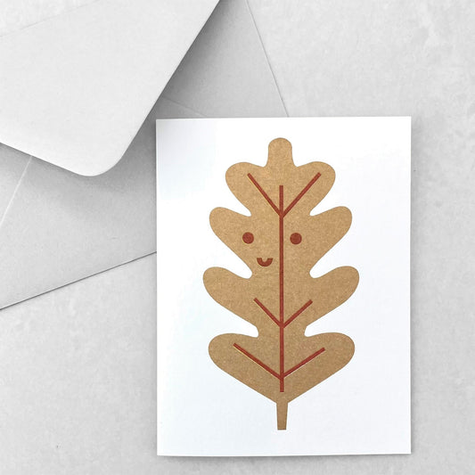 Mini greetings card with an image of a gold oak leaf with a smily face. By Scout Editions.