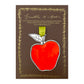 an apple shaped hanging ornament made of thick 1050gsm off-white card, letterpress printed in black and then hand painted in bright red ink. It has a chartreuse green satin ribbon to hang the ornament with. By Scribble & Daub. Pictured on a gold-foiled brown board.