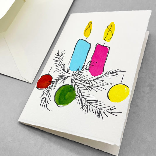 A letterpress printed card by Scribble & Daub. Two candles with foliage and baubles hand painted in a bright 70s colour palette of pink, red, yellow, light blue and green. Pictured with envelope