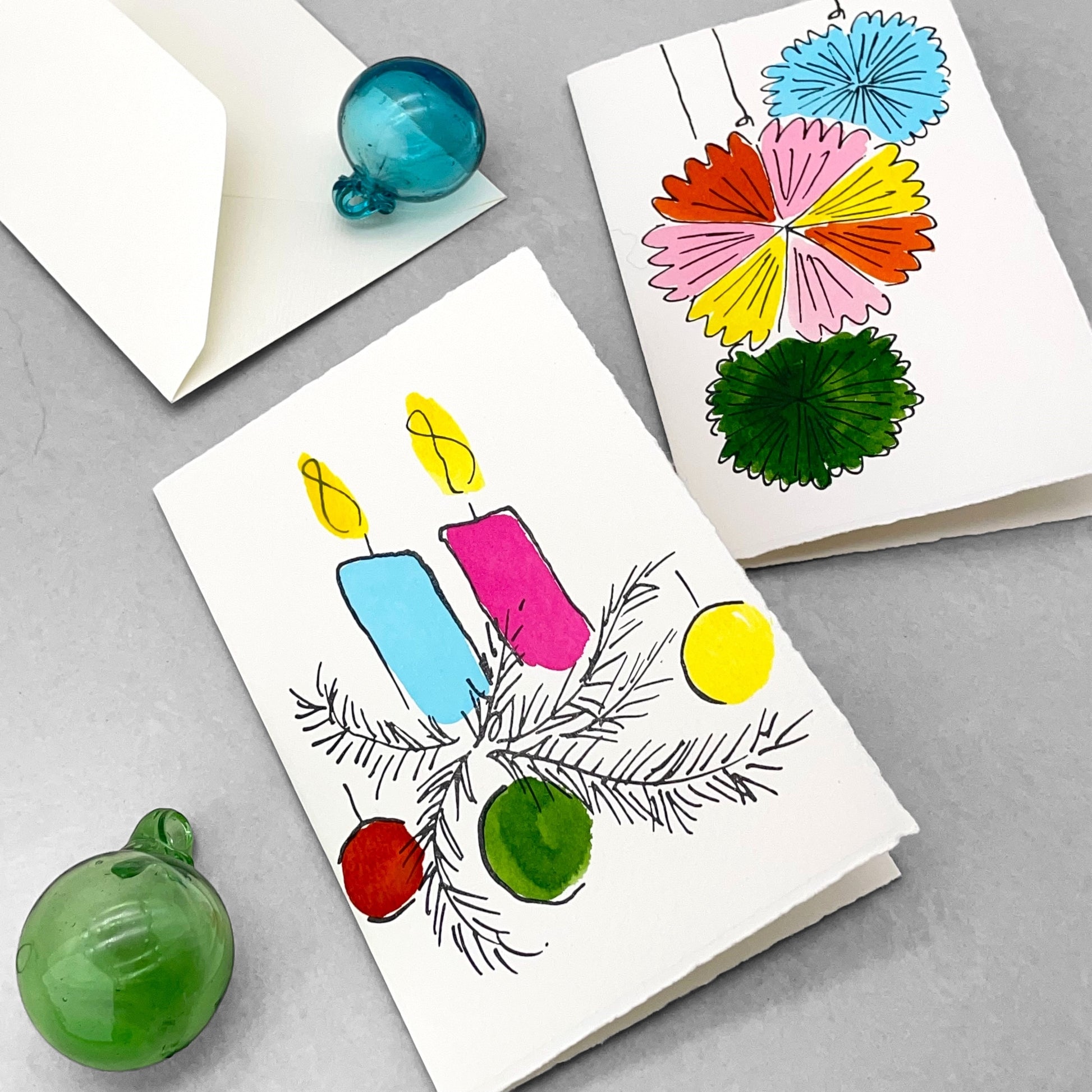 A letterpress printed card by Scribble & Daub. Two candles with foliage and baubles hand painted in a bright 70s colour palette of pink, red, yellow, light blue and green. Pictured with envelope, baubles and a pinwheel card from the same collection