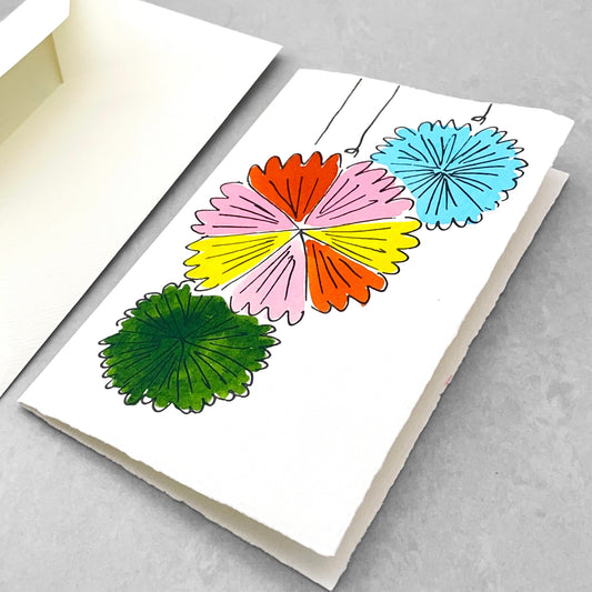 A letterpress printed card by Scribble & Daub. Three pinwheel decorations hand painted in a bright 70s colour palette of pink, red, yellow, light blue and green. Pictured with envelope