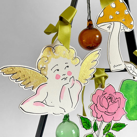 a cherub shaped hanging ornament made of thick 1050gsm off-white card, letterpress printed in black and then hand painted in pale pink and gold ink. It has a chartreuse green satin ribbon to hang the ornament with. By Scribble & Daub. Pictured hanging with baubles and other decorations from Scribble & Daub