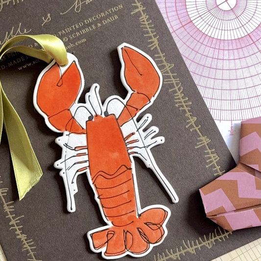 a lobster shaped hanging ornament made of thick 1050gsm off-white card, letterpress printed in black and then hand painted in orange ink. It has a chartreuse green satin ribbon to hang the ornament with. By Scribble & Daub. Pictured  next to a heart