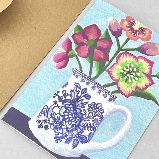 greeting card of a painting of purple hellebores in a blue and white patterned jug, with a pale blue background by Susie Hamilton