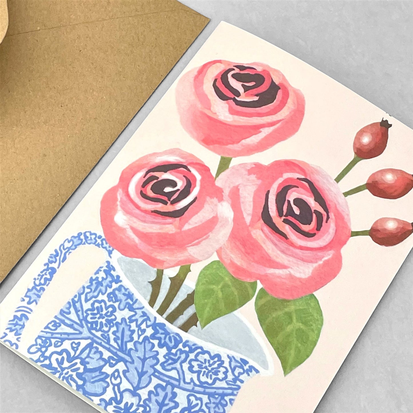 greeting card of a painting of pink roses in a blue and white chintz jug, pale blush pink backdrop by Susie Hamilton