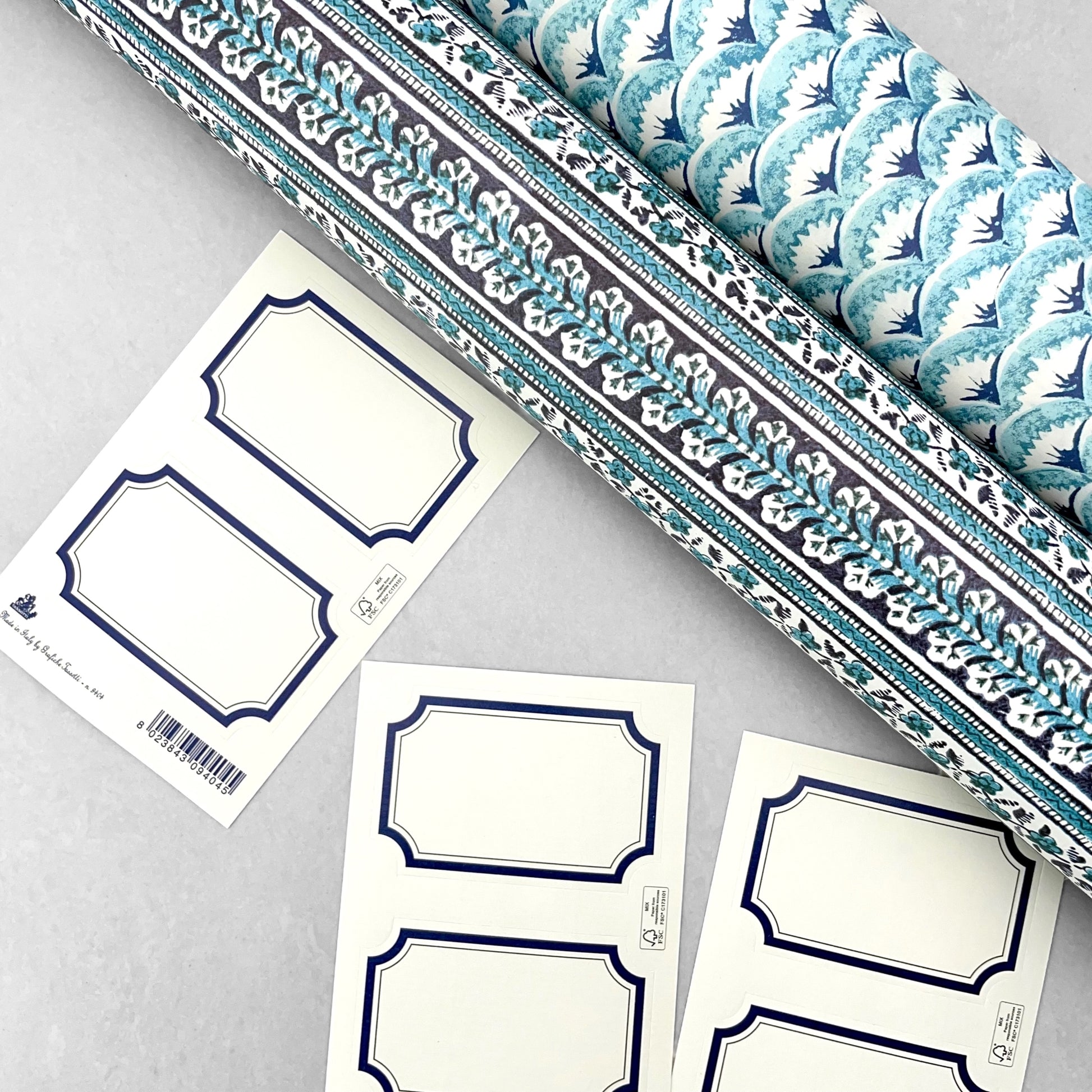 Adhesive labels made of ivory paper with a blue rectangular border by Grafiche Tassotti. Pictured alongside rolled wrapping paper