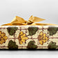 italian Remondini patterned wrapping paper by Tassotti. Repeat floral pattern with yellow and green flowers on a beige background with script writing. Close up of a present wrapped in the paper with a gold ribbon bow