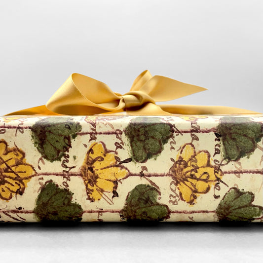 italian Remondini patterned wrapping paper by Tassotti. Repeat floral pattern with yellow and green flowers on a beige background with script writing. Close up of a present wrapped in the paper with a gold ribbon bow