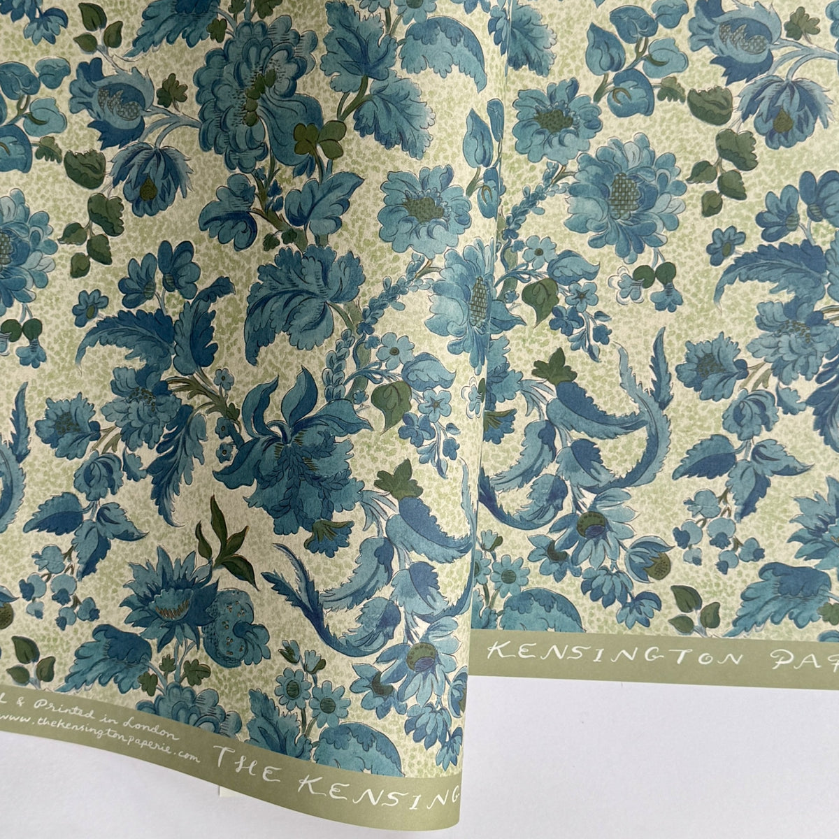 a sheet of wrapping paper by The Kensington Paperie with a large chintz floral in teal blue, with green foliage and a speckled light green ground 