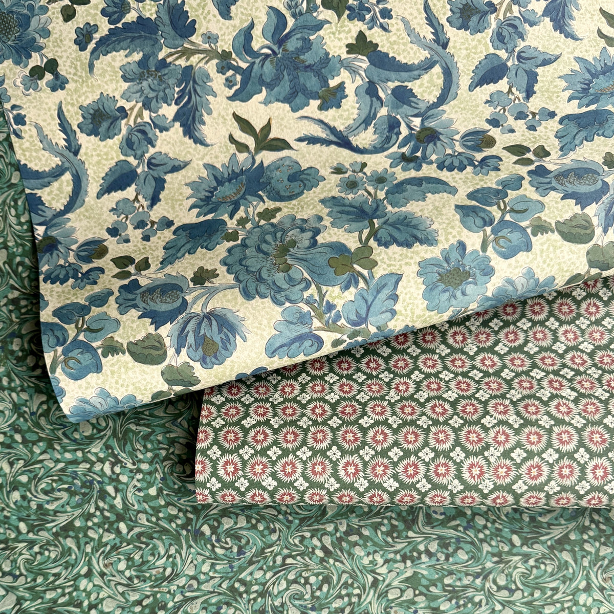 a sheet of wrapping paper by The Kensington Paperie with a large chintz floral in teal blue, with green foliage and a speckled light green ground.  Pictured alongside a geometric and a marbled paper 