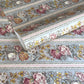 A floral patterned wrapping paper with large flowers in pink, cream, yellow, and muted green on a pale blue-gray background, divided by narrow decorative borders.