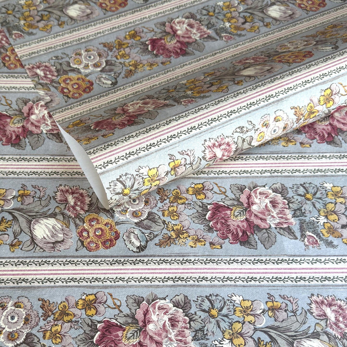 A floral patterned wrapping paper with large flowers in pink, cream, yellow, and muted green on a pale blue-gray background, divided by narrow decorative borders.