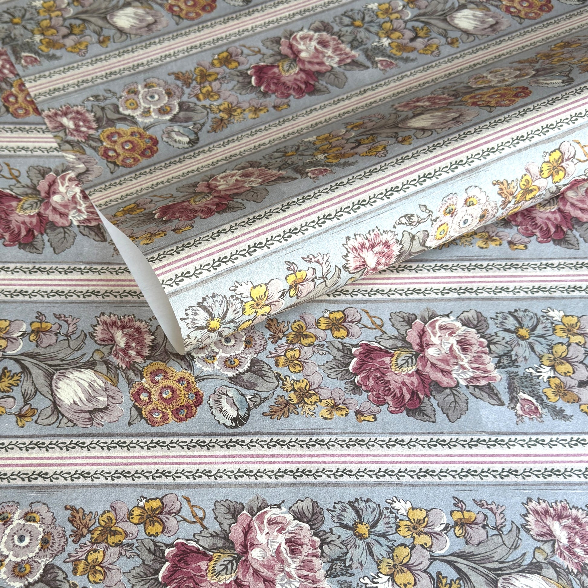 A floral patterned wrapping paper with large flowers in pink, cream, yellow, and muted green on a pale blue-gray background, divided by narrow decorative borders.