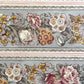 A close-up of a floral patterned wrapping paper with large flowers in pink, cream, yellow, and muted green on a pale blue-gray background, divided by narrow decorative borders.
