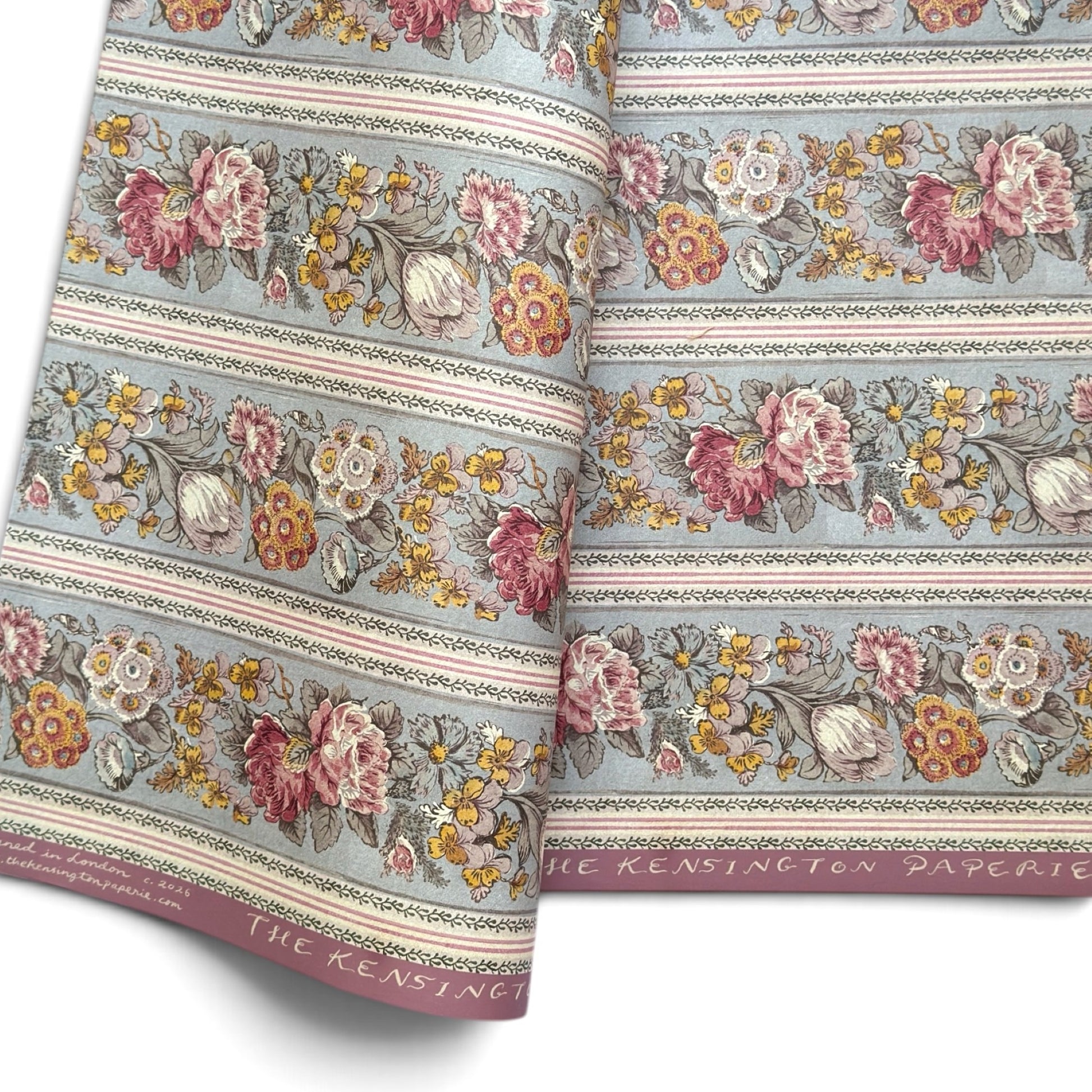 A floral patterned wrapping paper with large flowers in pink, cream, yellow, and muted green on a pale blue-gray background, divided by narrow decorative borders.