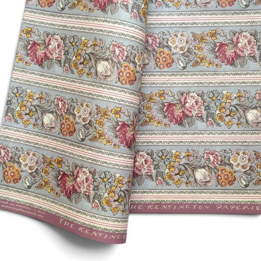 A floral patterned wrapping paper with large flowers in pink, cream, yellow, and muted green on a pale blue-gray background, divided by narrow decorative borders.