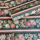 A sheet of wrapping paper, showing a detailed pattern of pink, white, and yellow flowers with green leaves on a black, bordered by horizontal stripes in pink, white, and brown.