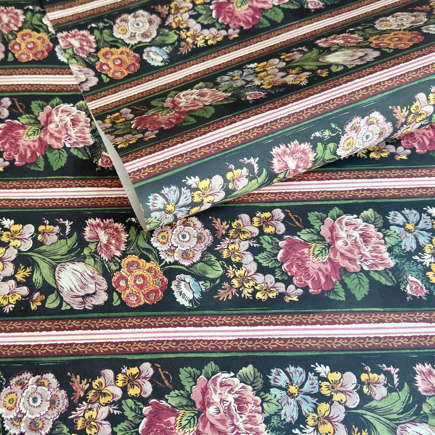 A sheet of wrapping paper, showing a detailed pattern of pink, white, and yellow flowers with green leaves on a black, bordered by horizontal stripes in pink, white, and brown.