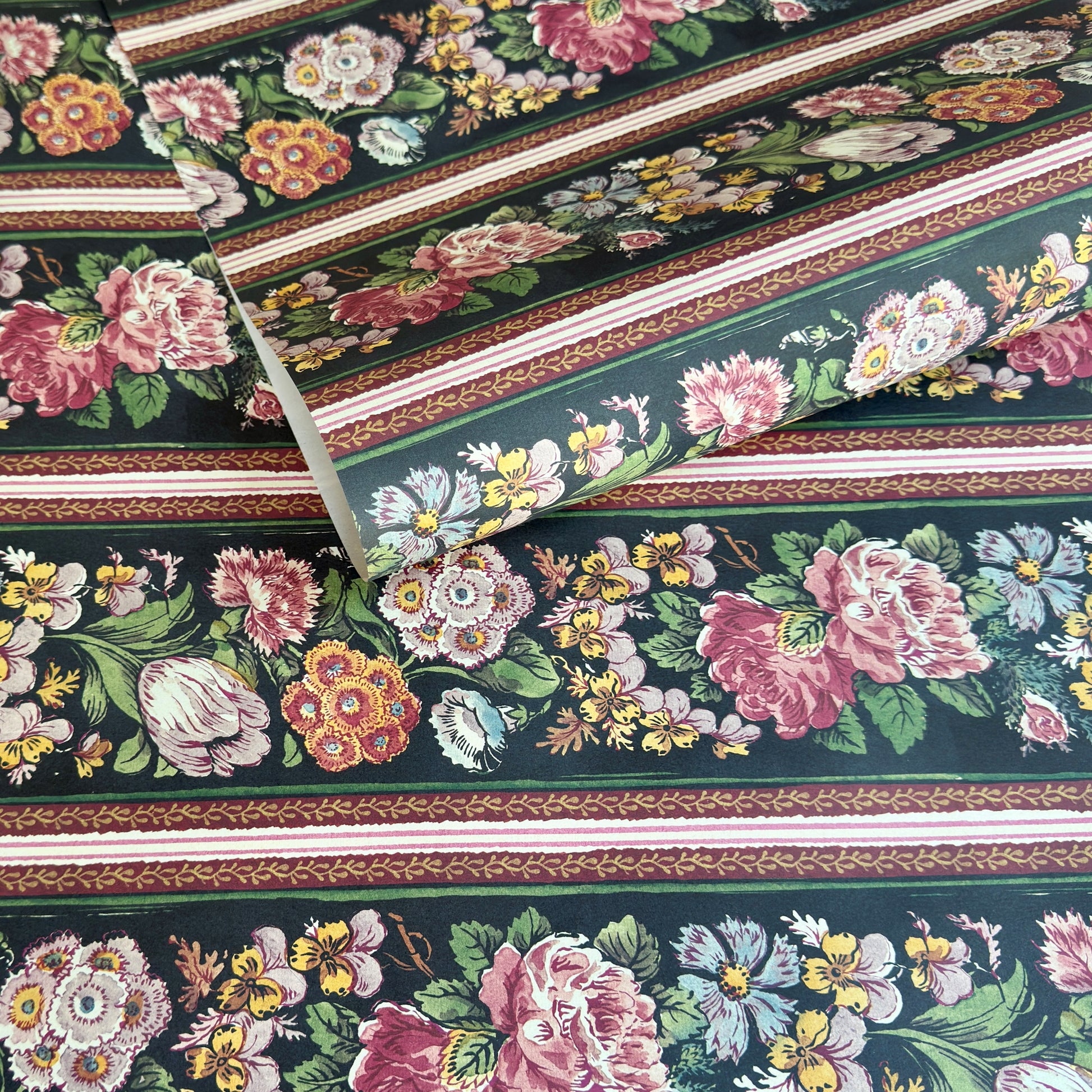 A sheet of wrapping paper, showing a detailed pattern of pink, white, and yellow flowers with green leaves on a black, bordered by horizontal stripes in pink, white, and brown.