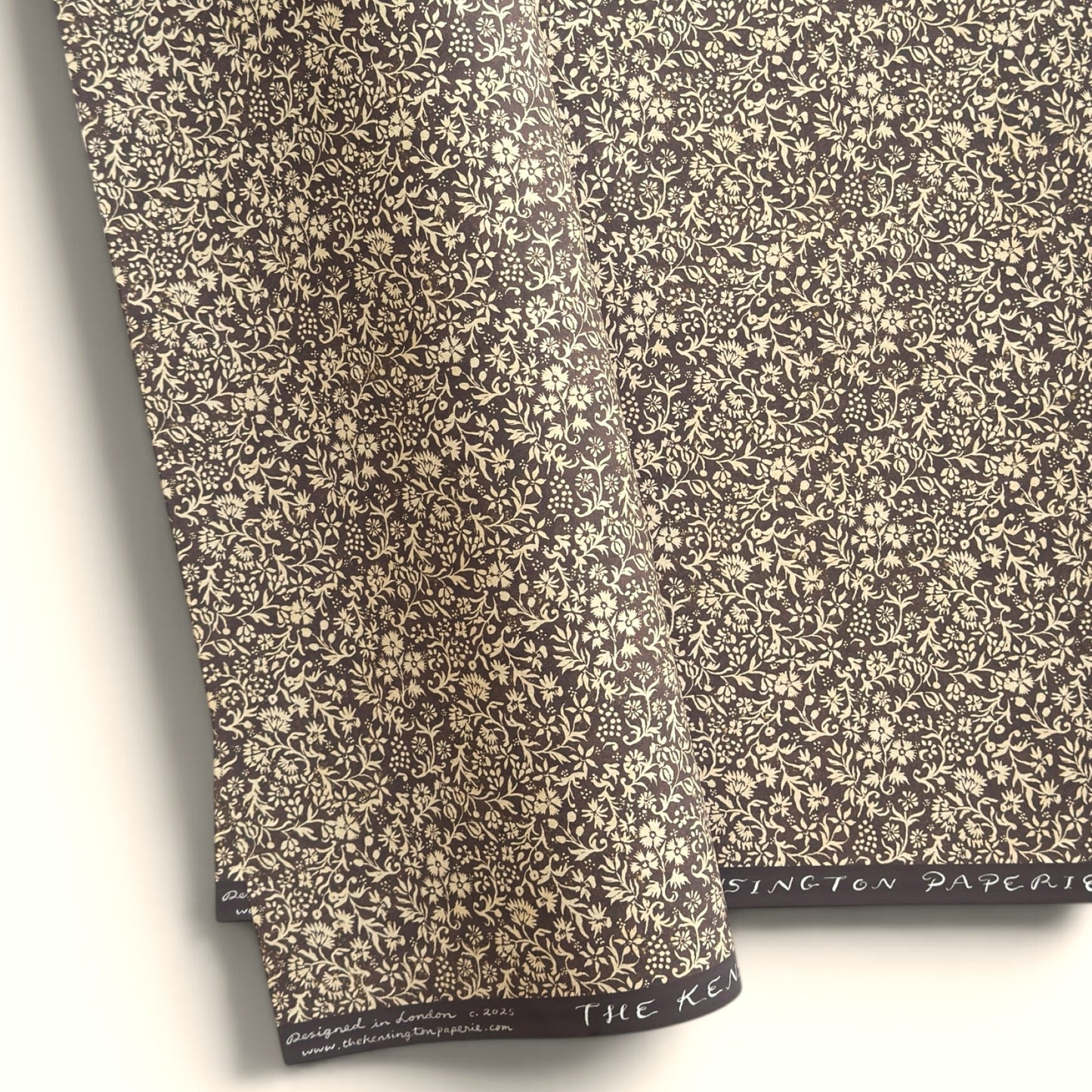 A sheet of dark aubergine wrapping paper with a dense cream floral and vine pattern, shown partially unrolled on a white background. 