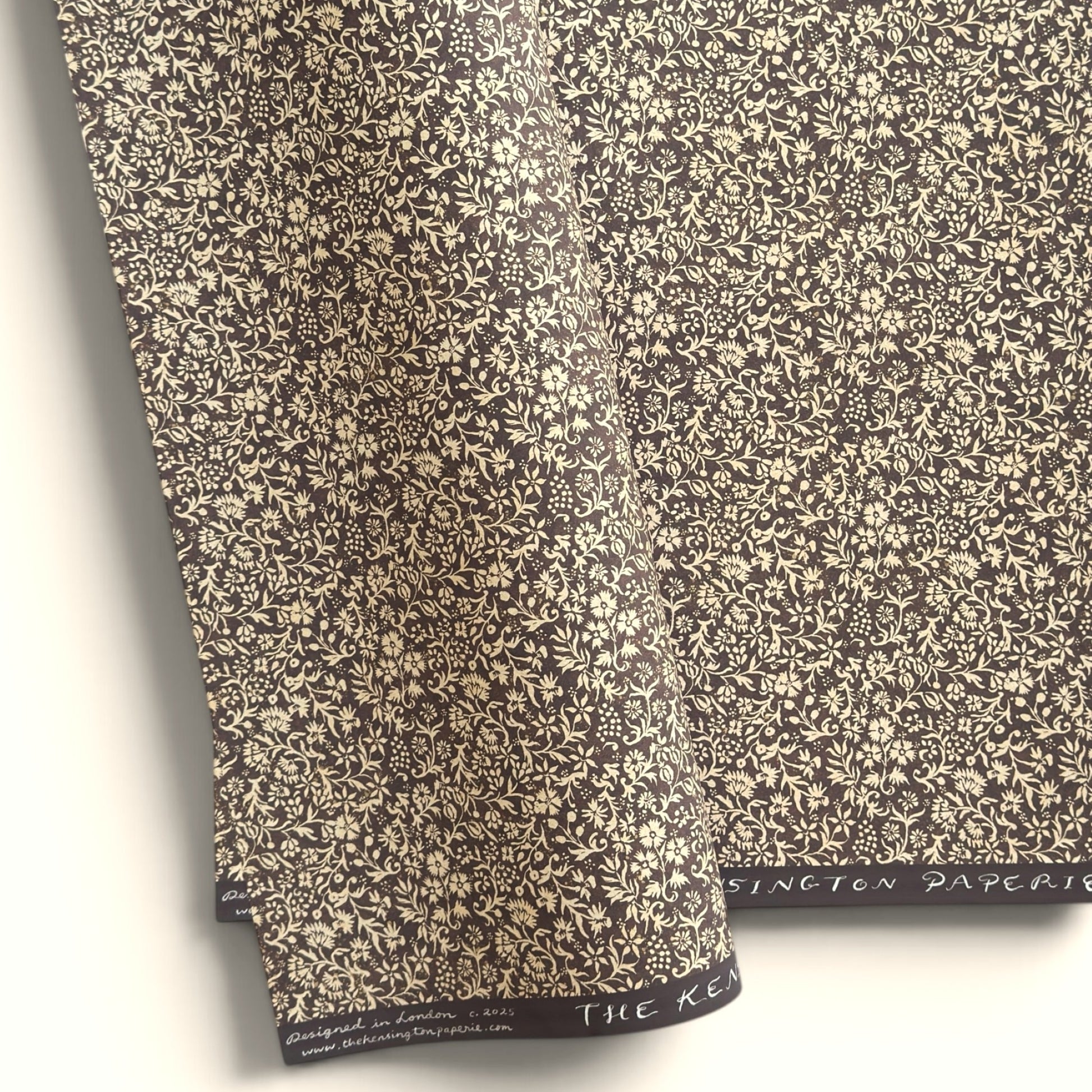 A sheet of dark aubergine wrapping paper with a dense cream floral and vine pattern, shown partially unrolled on a white background. 