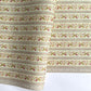 A close-up of patterned wrapping paper with horizontal stripes and a repeating floral vine motif in muted green, pink, and brown on an off-white background. A band at the bottom reads 'THE KENSINGTON PAPER' with smaller text below.