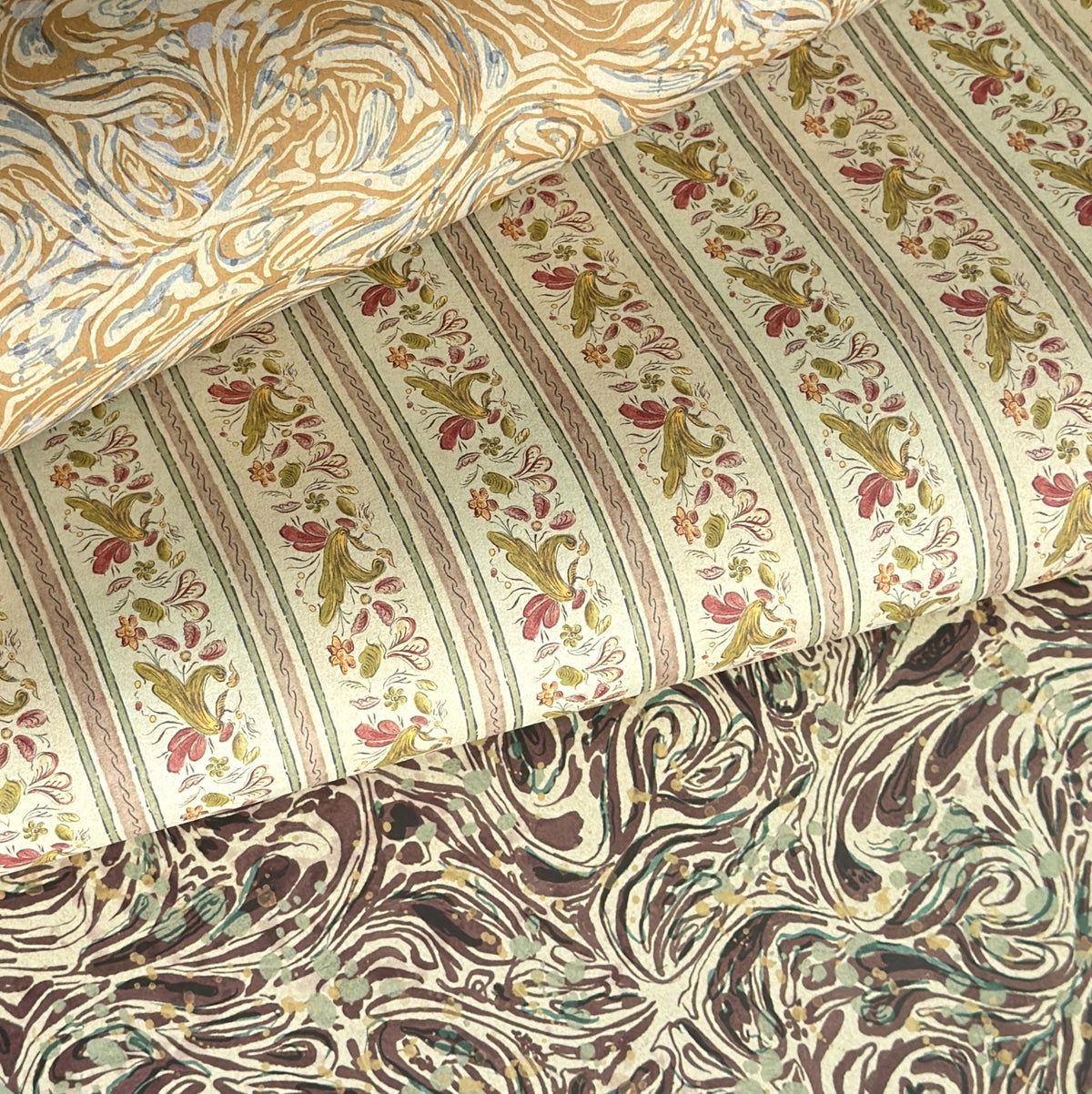 a close up of three sheets of patterned wrapping paper, top and bottom sheets are marbled designs is sandy and brown colours. Middle sheet is a stripe with a floral motif