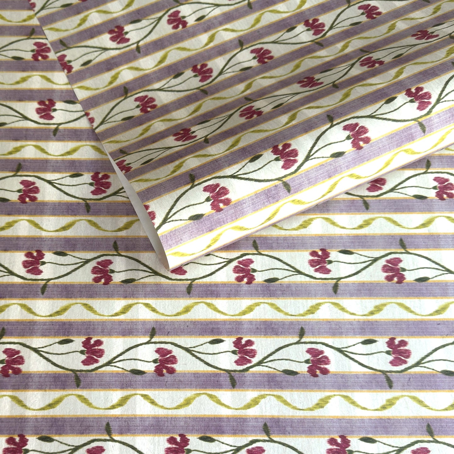 A close-up of a patterned wrapping paper featuring horizontal lavender and cream stripes with pink flowers, green stems, and olive-green wavy lines.