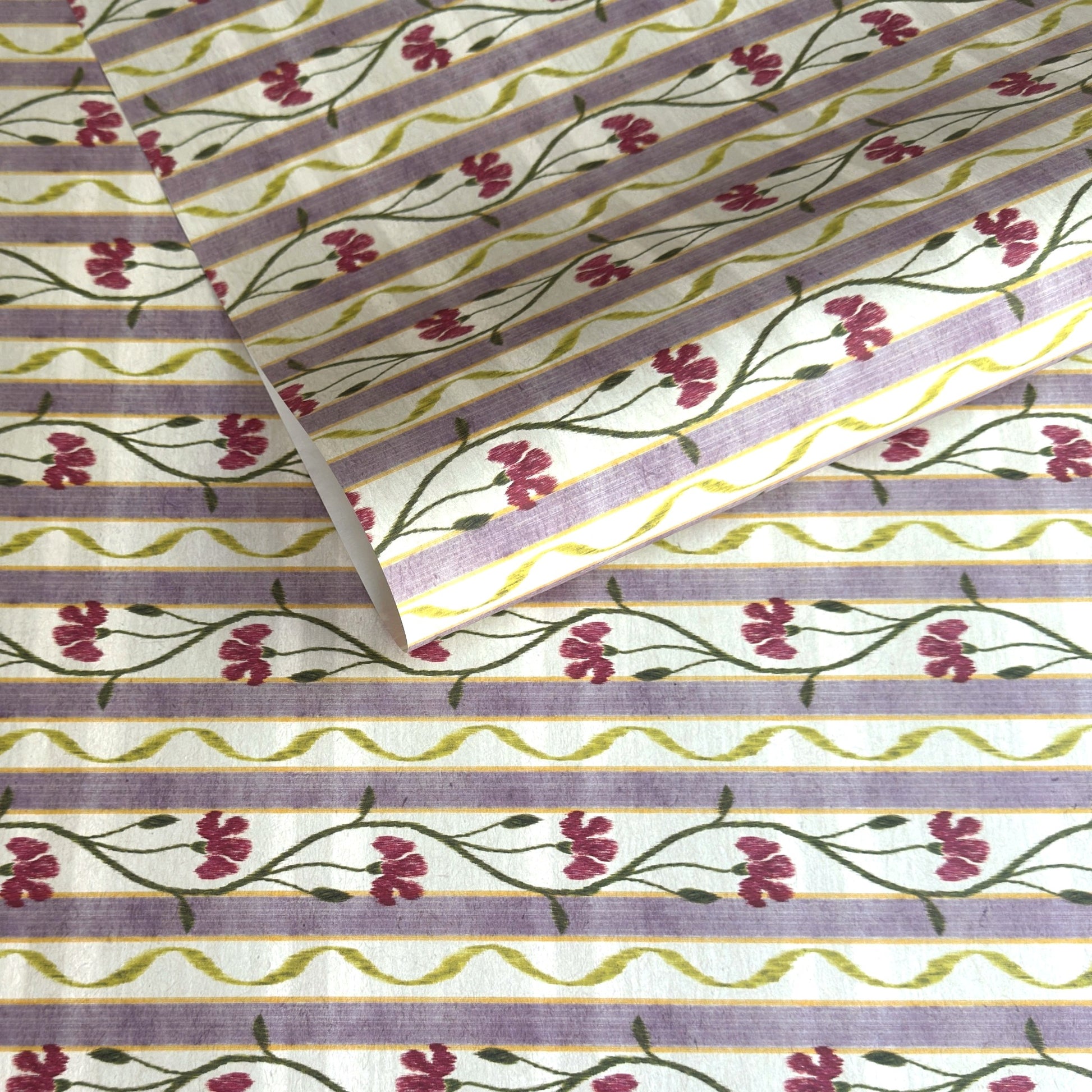 A close-up of a patterned wrapping paper featuring horizontal lavender and cream stripes with pink flowers, green stems, and olive-green wavy lines.