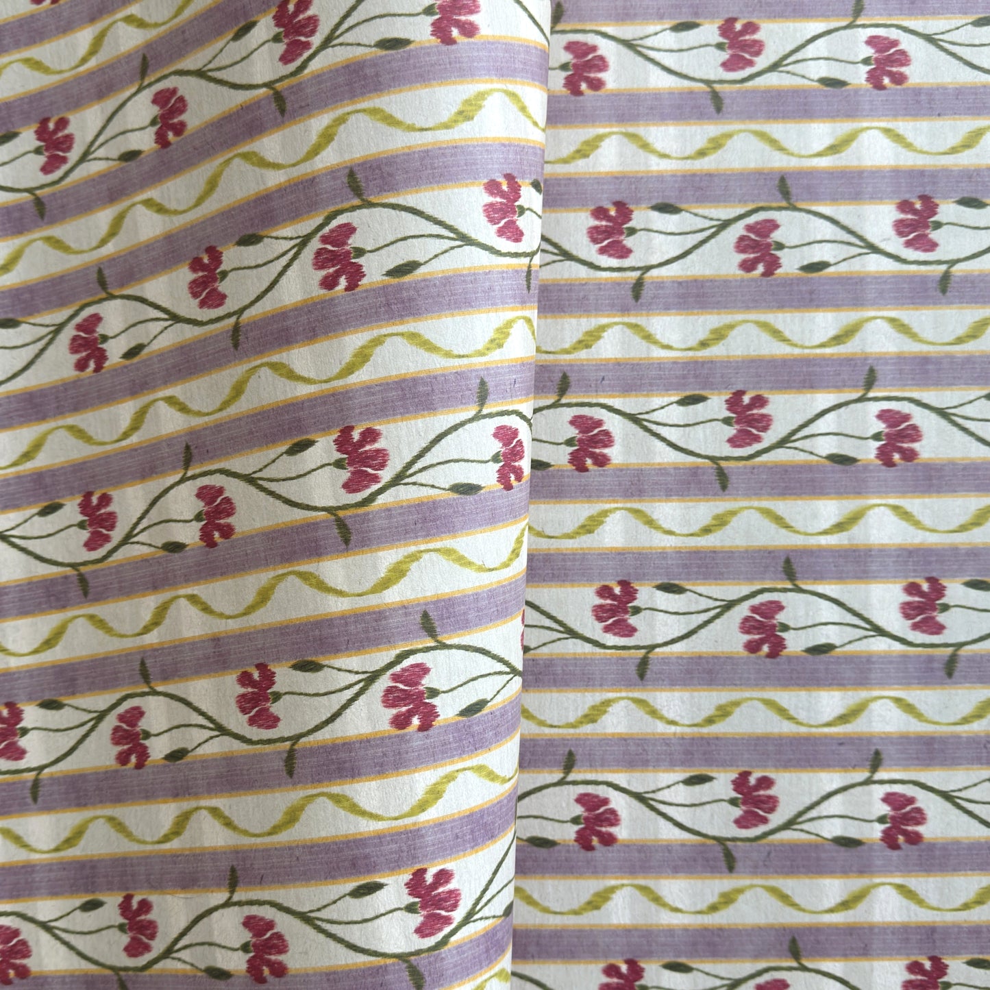 A close-up of a patterned wrapping paper featuring horizontal lavender and cream stripes with pink flowers, green stems, and olive-green wavy lines.
