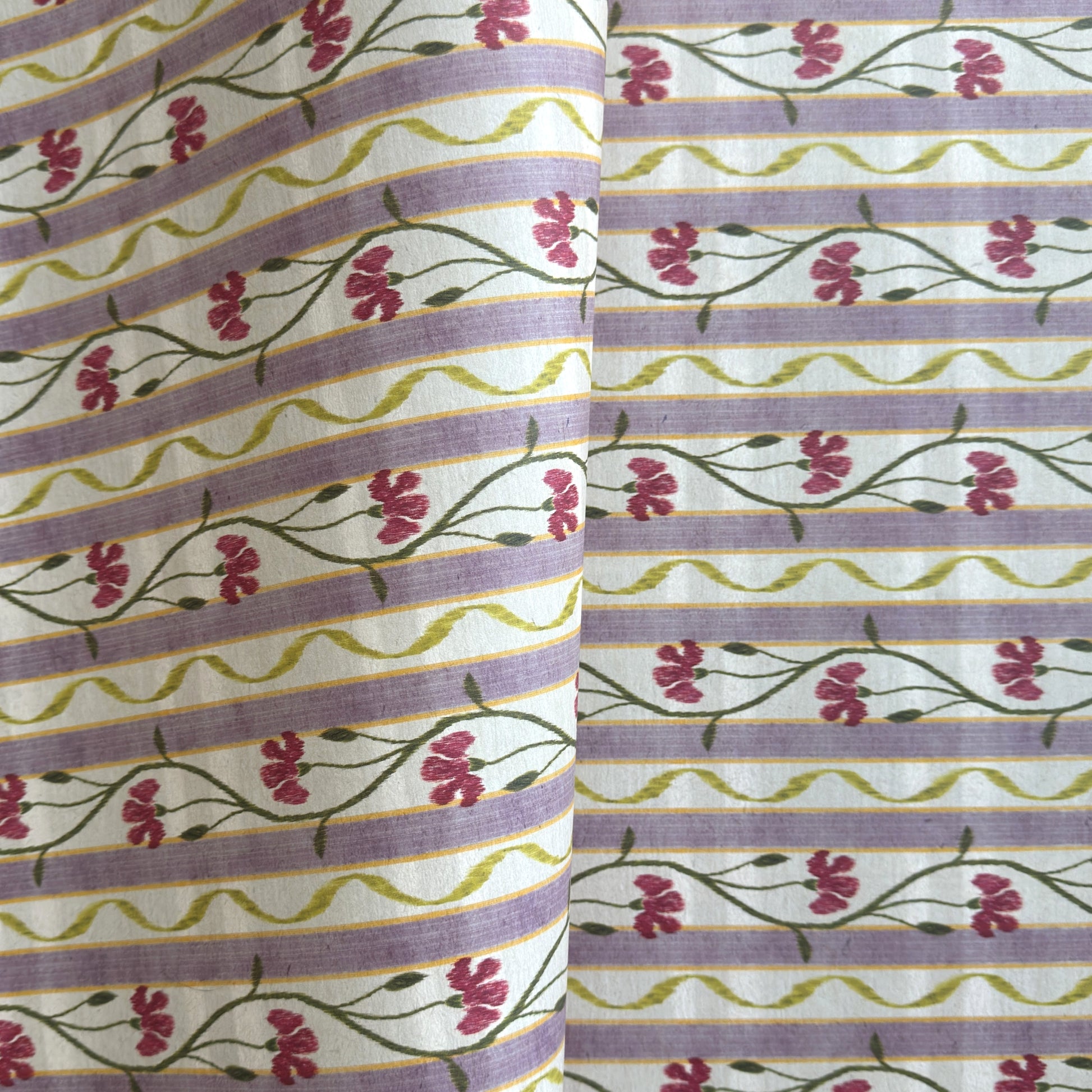A close-up of a patterned wrapping paper featuring horizontal lavender and cream stripes with pink flowers, green stems, and olive-green wavy lines.
