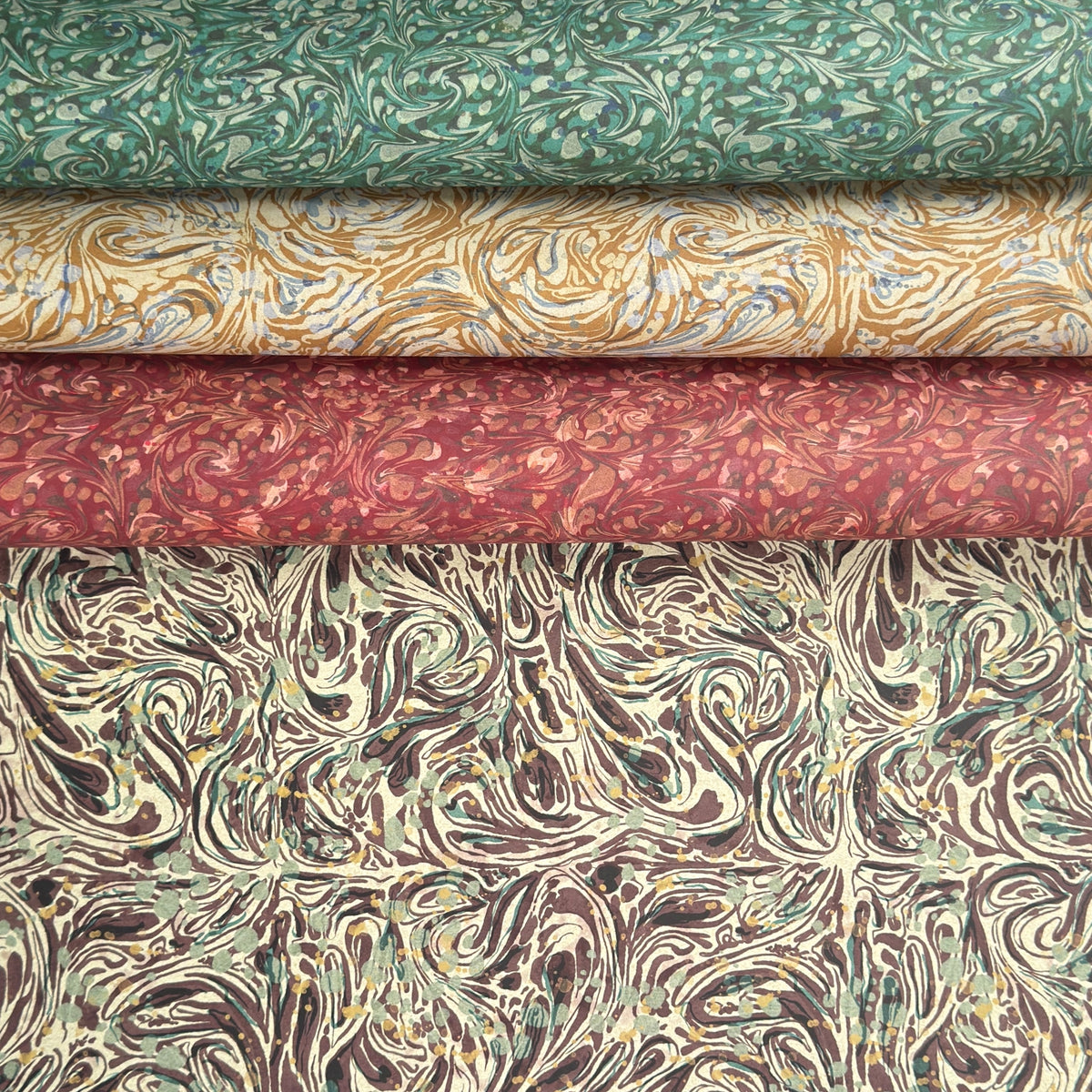 a printed marbled wrapping paper sheet with swirling brown, beige, muted green, and small yellow accents. By The Kensington Paperie brand. Pictured alongside versions in red, yellow and green