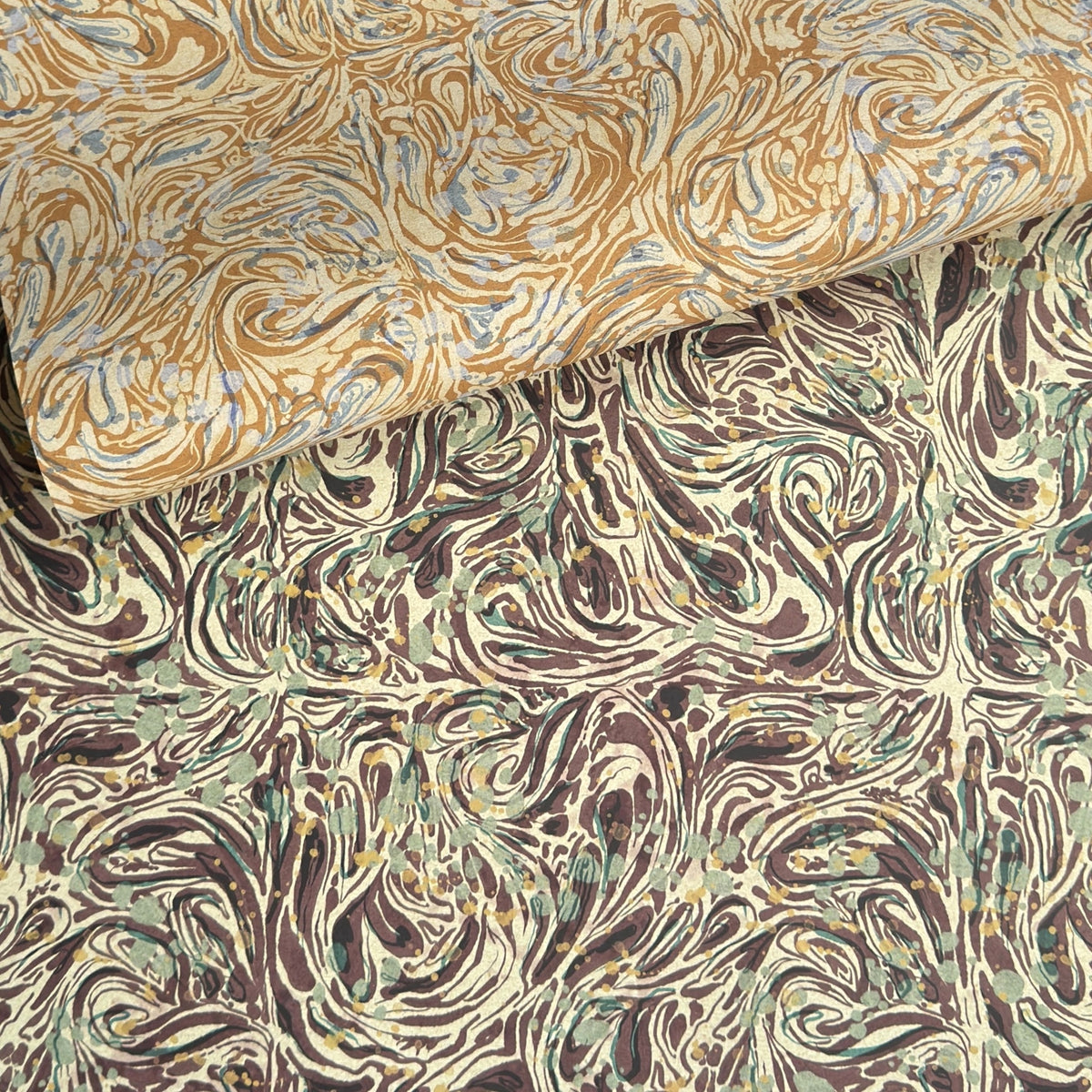 a printed marbled wrapping paper sheet with swirling brown, beige, muted green, and small yellow accents. By The Kensington Paperie brand. Pictured alongside a version in yellow