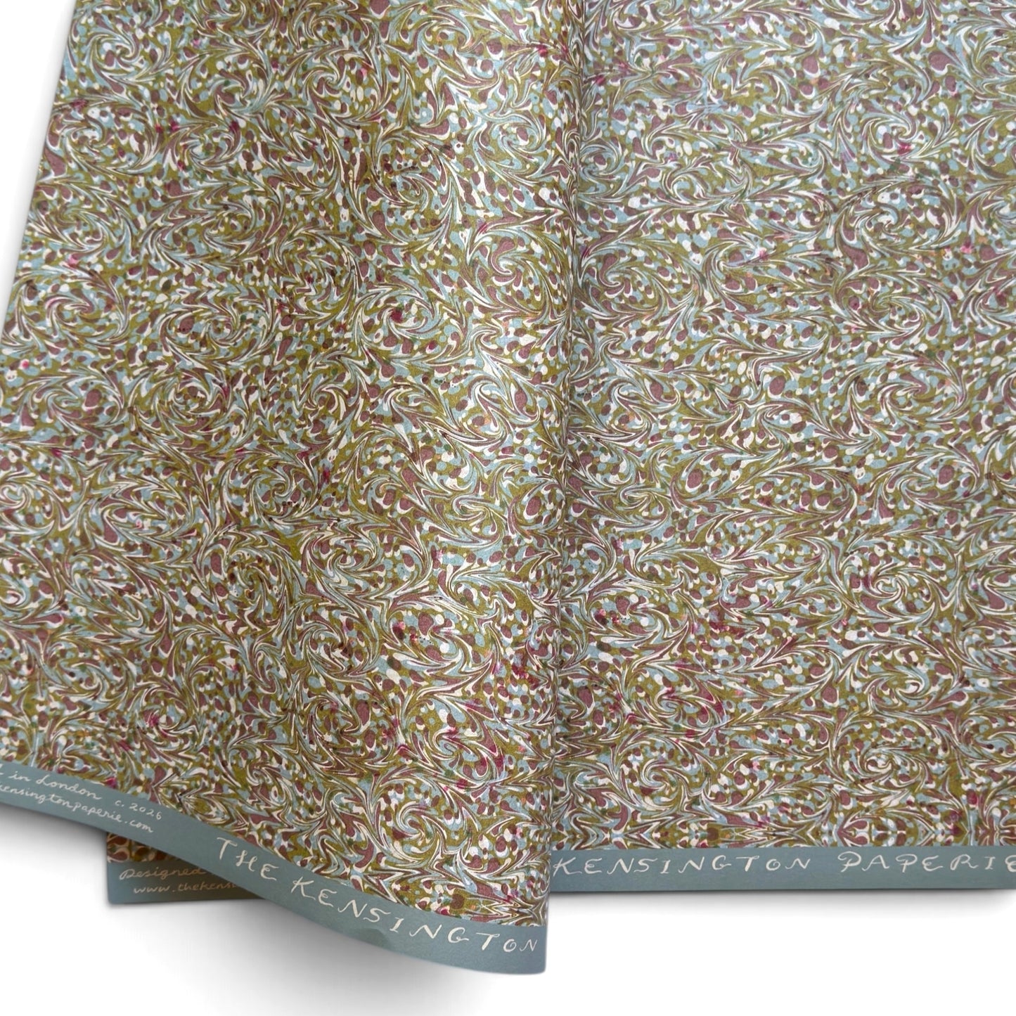 A folded roll of patterned paper with an intricate swirling floral design in muted gold, dusty rose, olive green, and white. The paper is shown folded to display both the patterned surface and the branded paper edge, which reads 'THE KENSINGTON PAPERIE'.