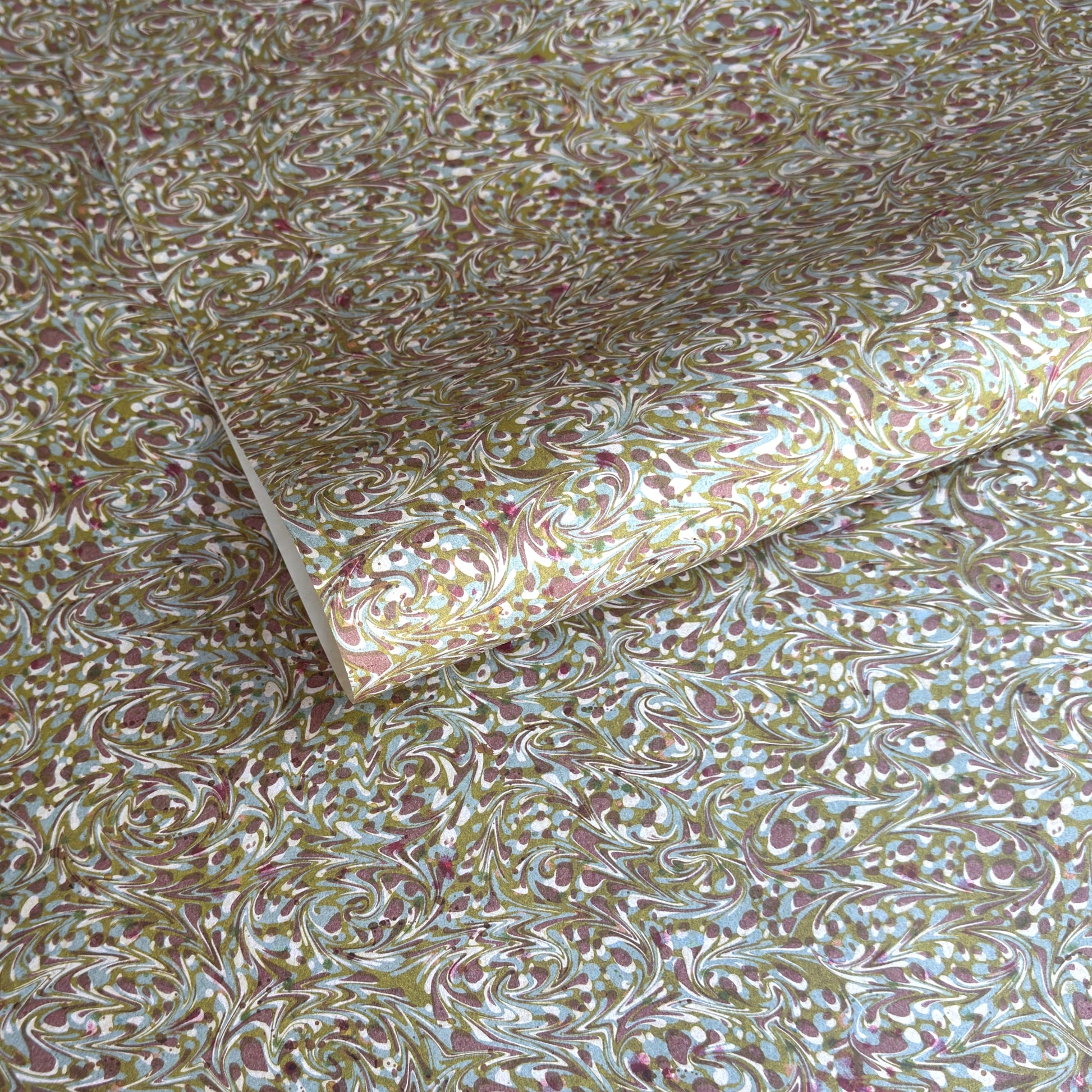 A close-up of patterned paper with an intricate marbled swirl design in olive green, gold, white, and dusty pink, with one corner folded over to show the paper's thickness.