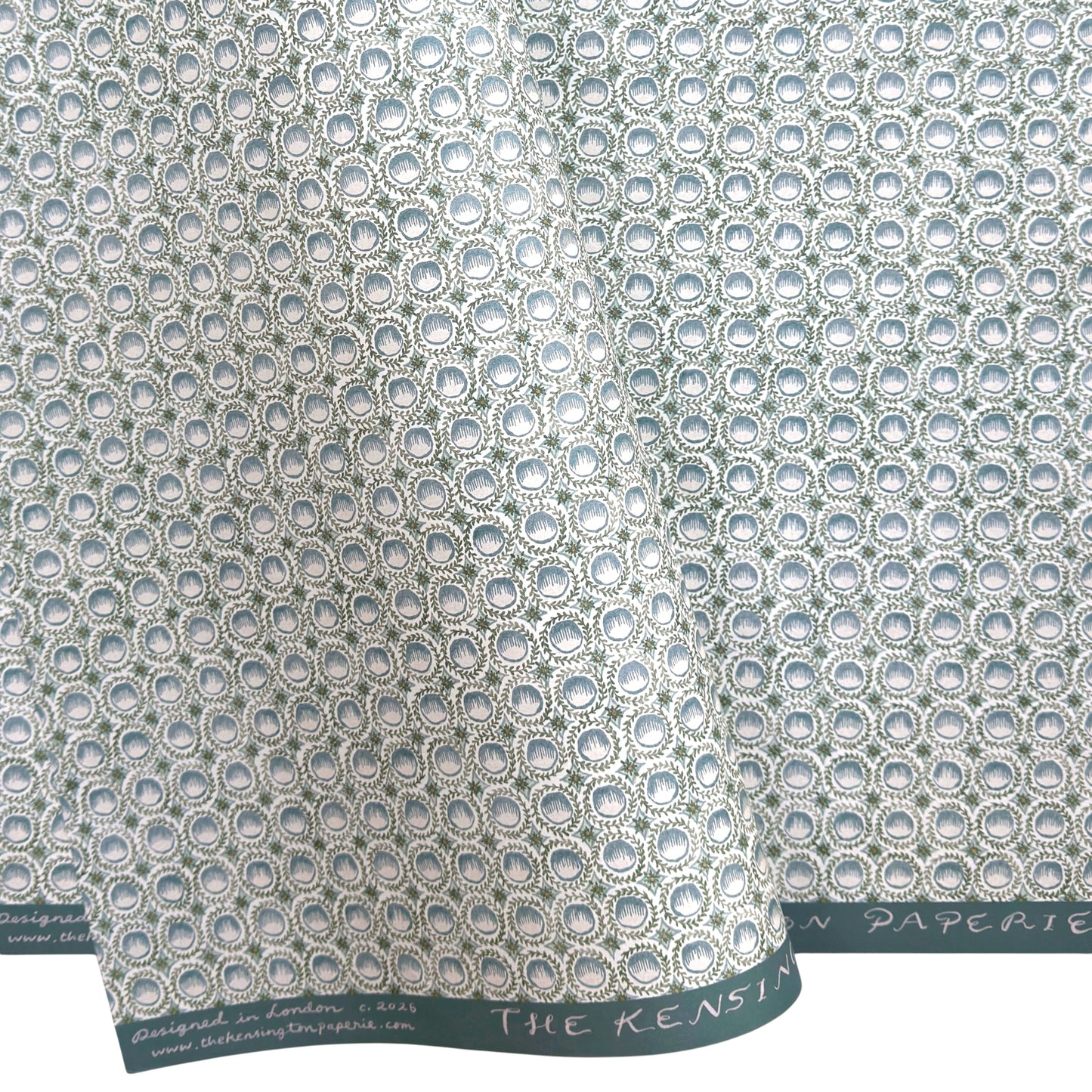 a patterned wrapping paper featuring a repeating botanical design with blue block printed steam puddings, green leafy wreaths, and small yellow accents on a white background.