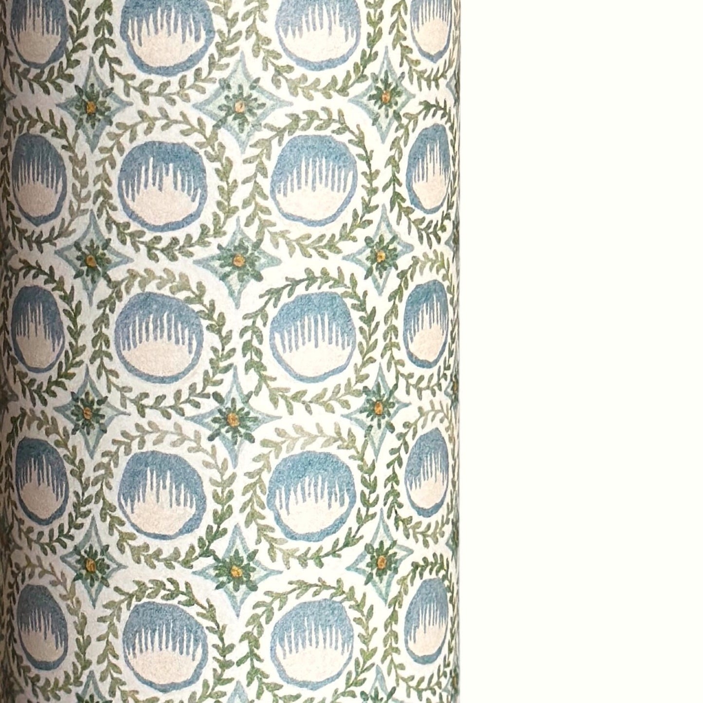 A close-up of a patterned wrapping paper featuring a repeating botanical design with blue block printed steam puddings, green leafy wreaths, and small yellow accents on a white background.