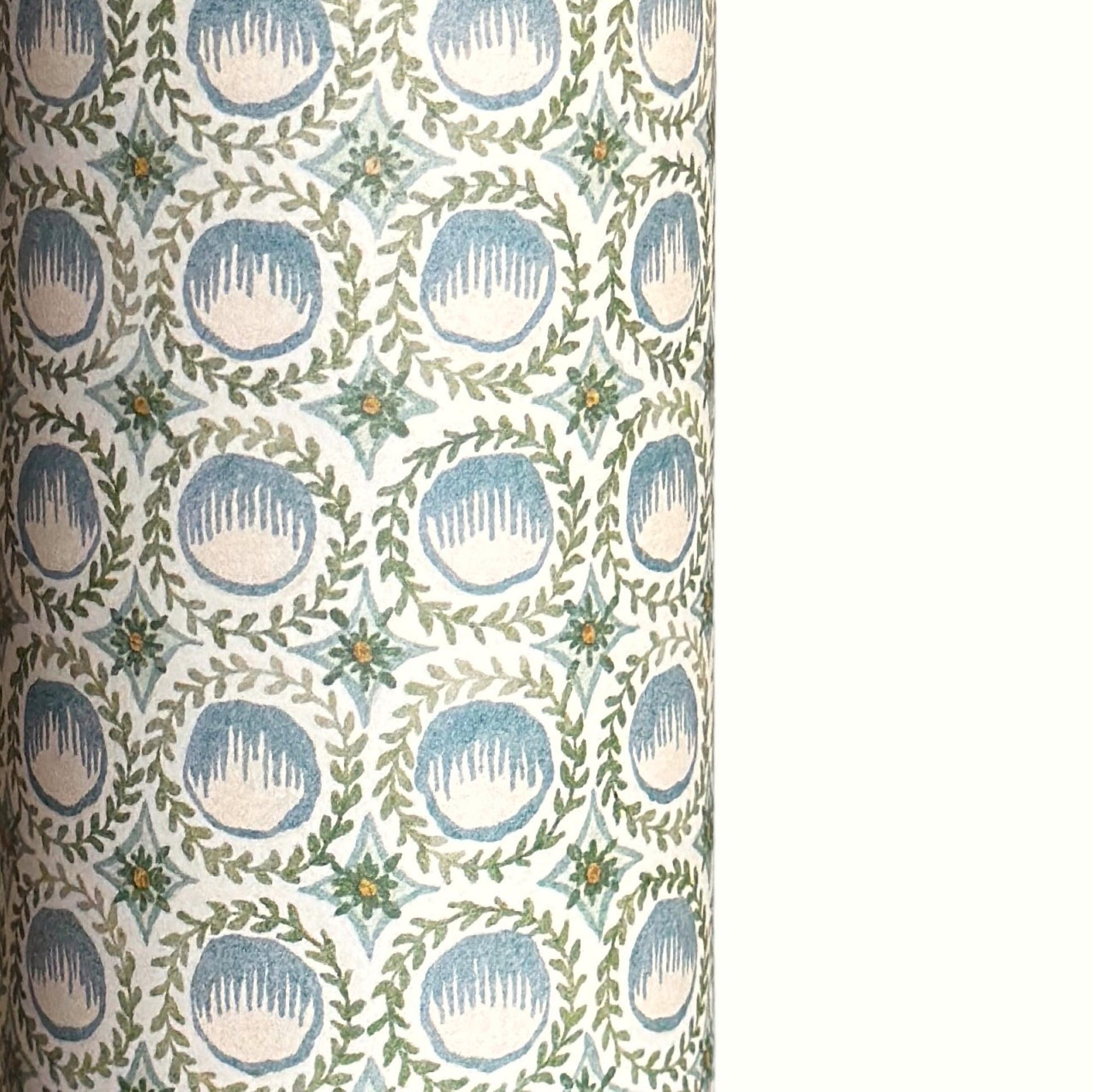 A close-up of a patterned wrapping paper featuring a repeating botanical design with blue block printed steam puddings, green leafy wreaths, and small yellow accents on a white background.