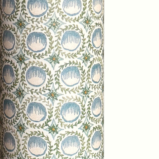 A close-up of a patterned wrapping paper featuring a repeating botanical design with blue block printed steam puddings, green leafy wreaths, and small yellow accents on a white background.