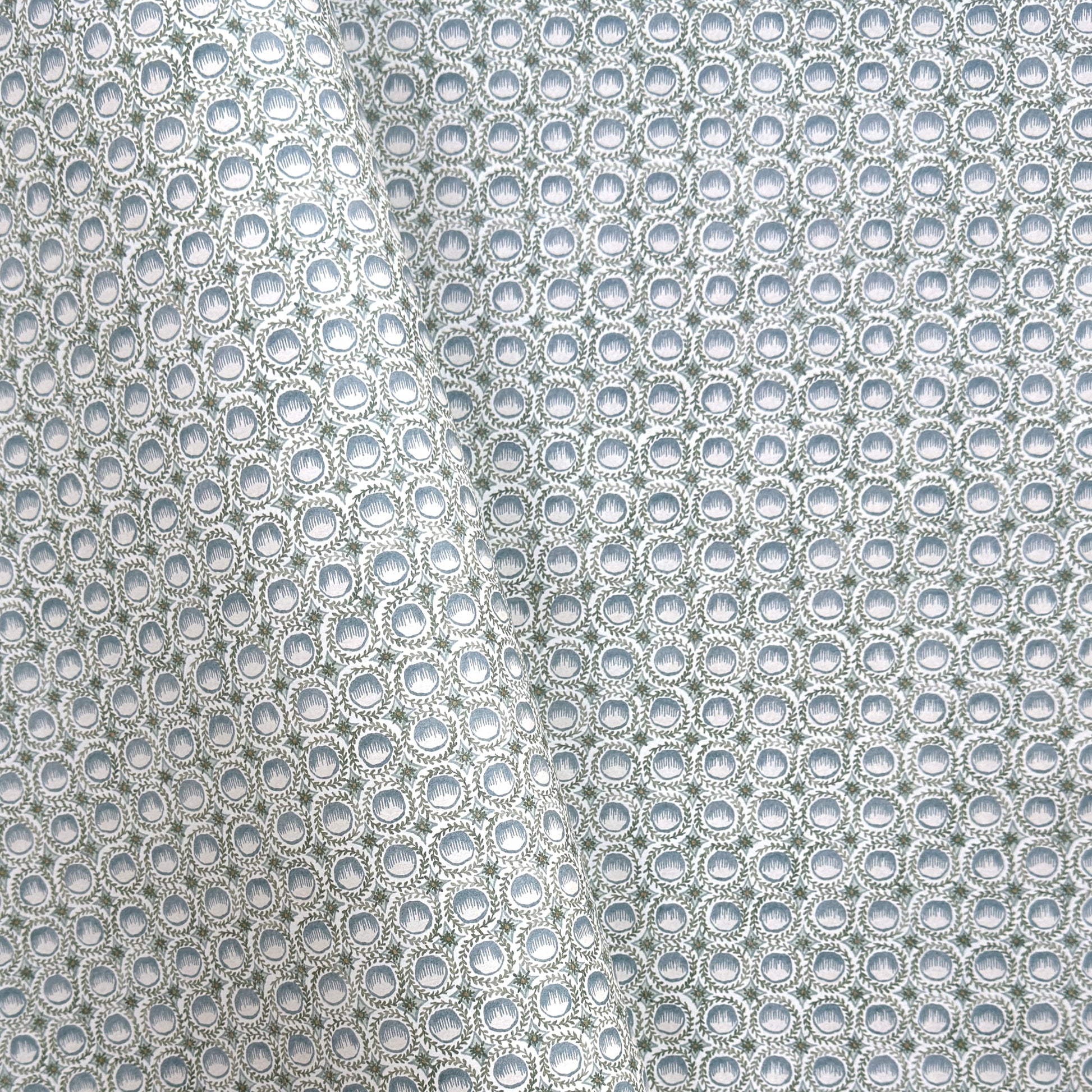 a patterned wrapping paper featuring a repeating botanical design with blue block printed steam puddings, green leafy wreaths, and small yellow accents on a white background.