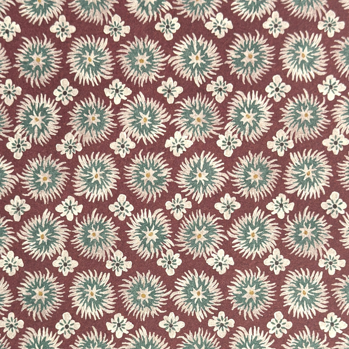 a sheet of wrapping paper by The Kensington Paperie with a small block printed geometric print in white and soft green on a cranberry ground. Close up