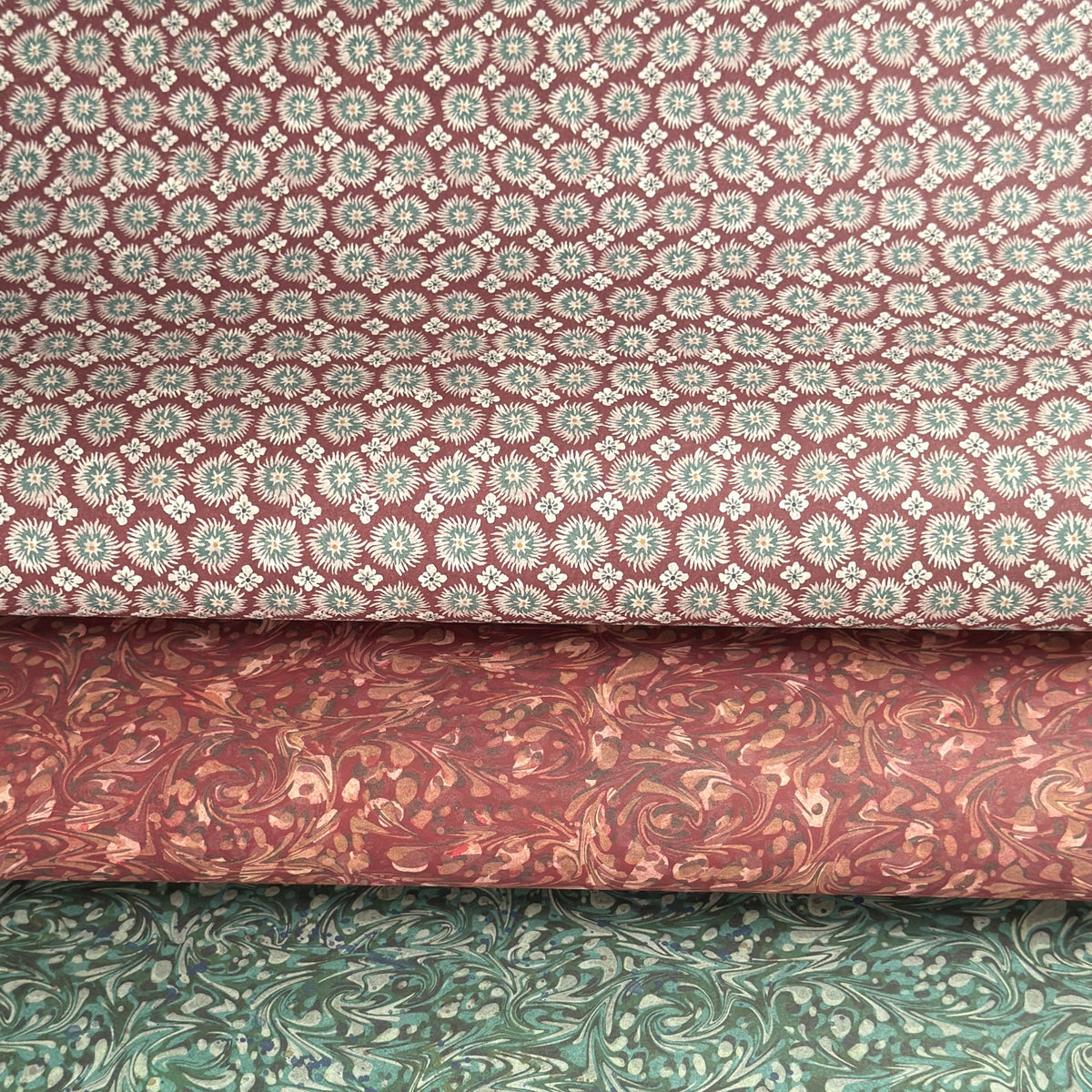 a sheet of wrapping paper by The Kensington Paperie with a small block printed geometric print in white and soft green on a cranberry ground.  Pictured rolled with a coordinating marble papers