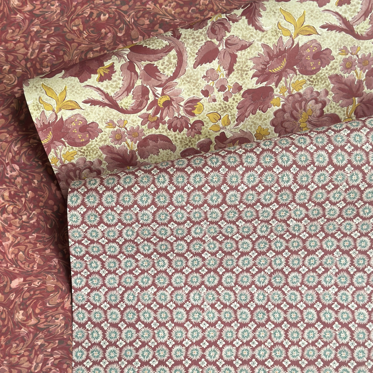 a sheet of wrapping paper by The Kensington Paperie with a small block printed geometric print in white and soft green on a cranberry ground. Pictured lightly folded with a complementary bold floral paper and a marbled design.