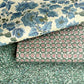 Three overlapping wrapping paper sheets with distinct patterns: a cream background with large blue floral motifs, a dark green background with repeating pink and white starburst medallions, and a teal-green background with an abstract swirling marbled pattern.