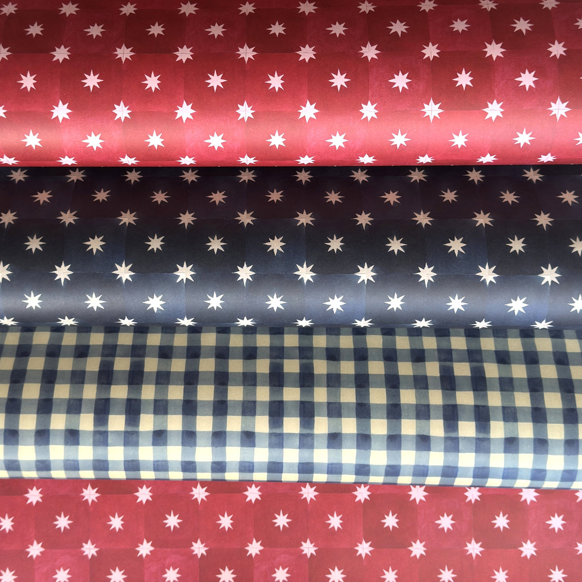 Red two-tone checked wrapping paper by Wanderlust with a white star pattern, shown lightly rolled with two other designs from the same collection, a navy star pattern and a blue gingham pattern