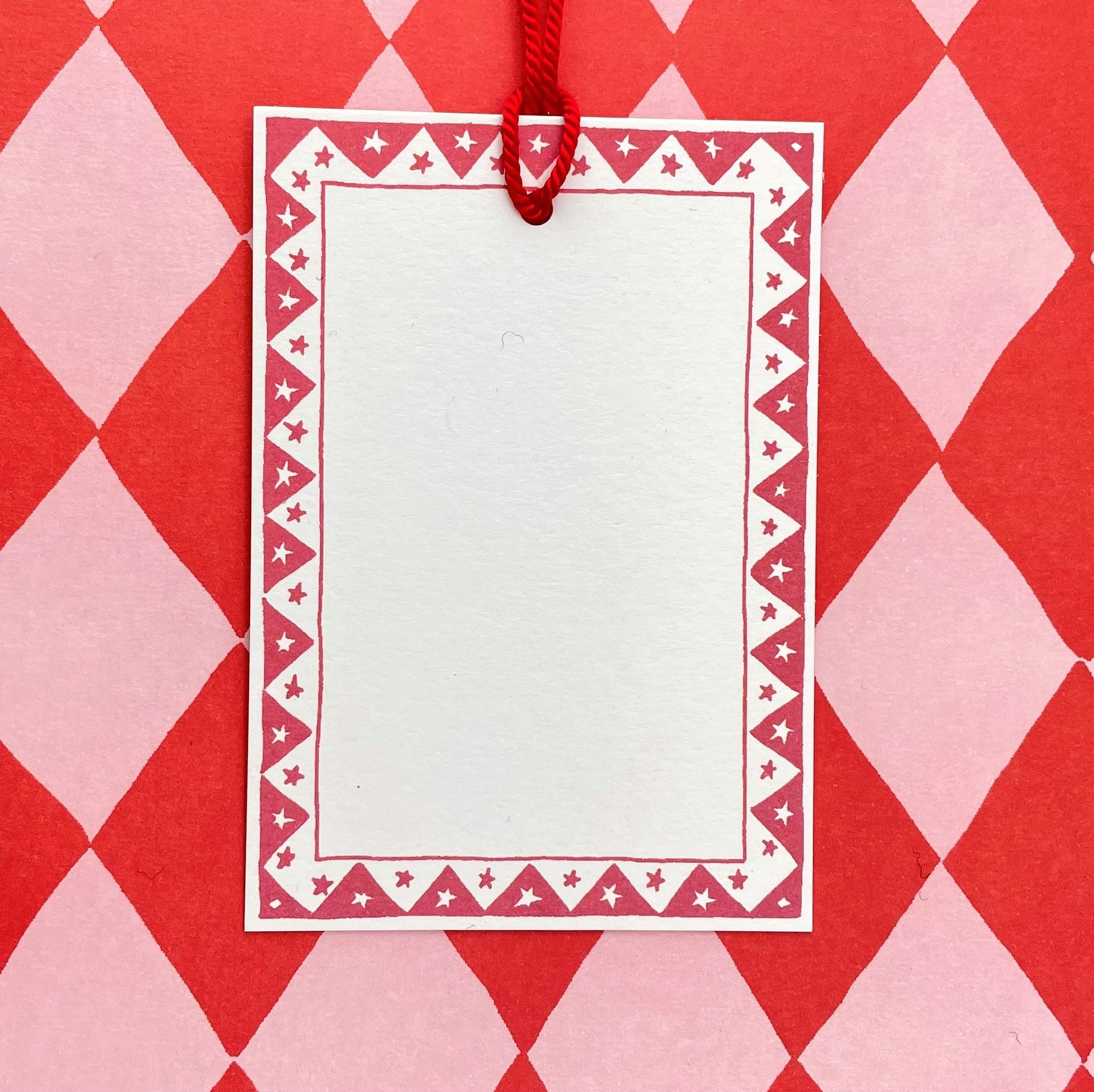 White rectangular gift tag with dark red star pattern border and red cord, pictured with red diamond wrapping paper