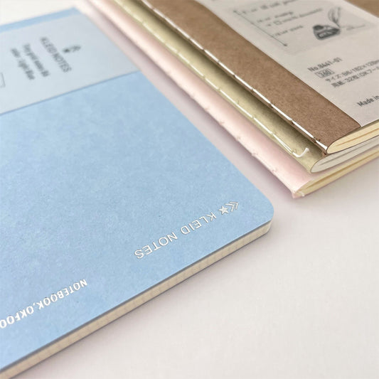 Soft cover notebook with grid format pages. Cover is plain light blue with branded belly band, close-up