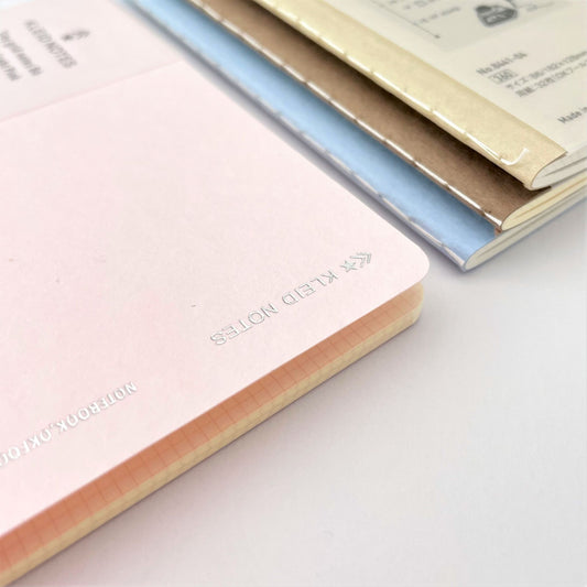 Soft cover notebook with grid format pages. Cover is plain light pink with branded belly band, close-up