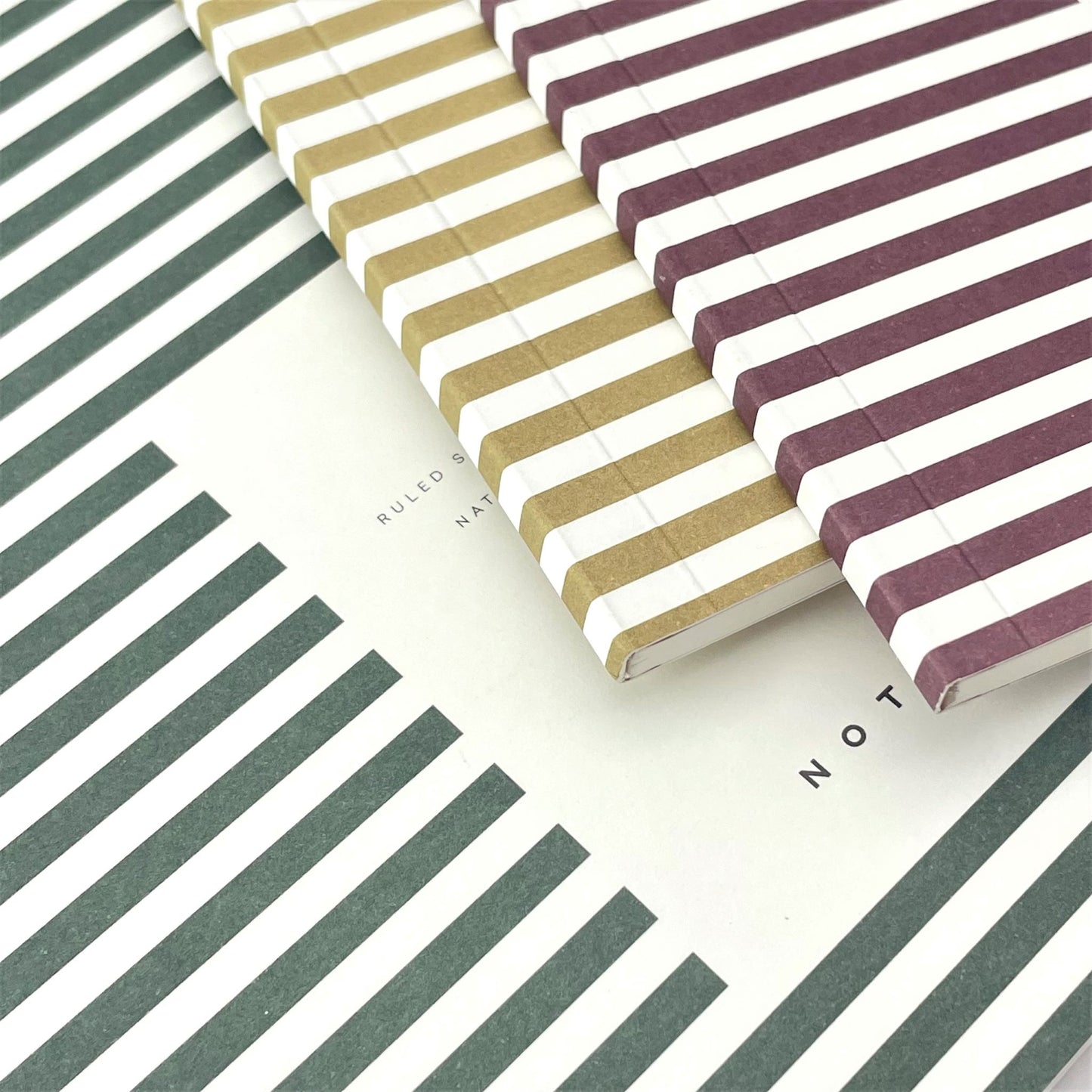 Pocket size notebook with a narrow dark green and white stripe softcover. pictured with other colour notebooks