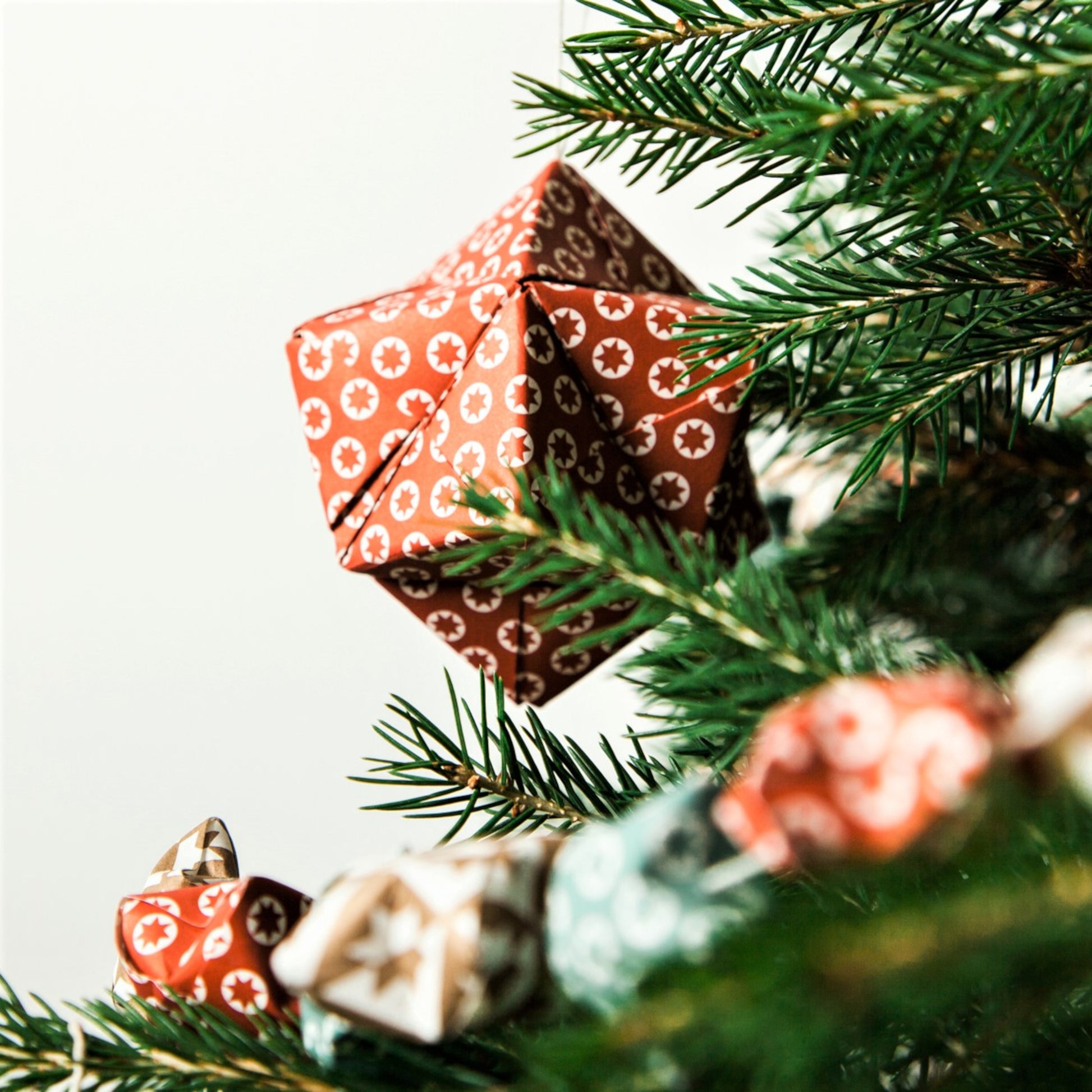 A set of patterned paper origami pyramid and bauble shaped decorations, in colours red, gold, navy and white, close-up of red paper bauble hung on a christmas tree branch